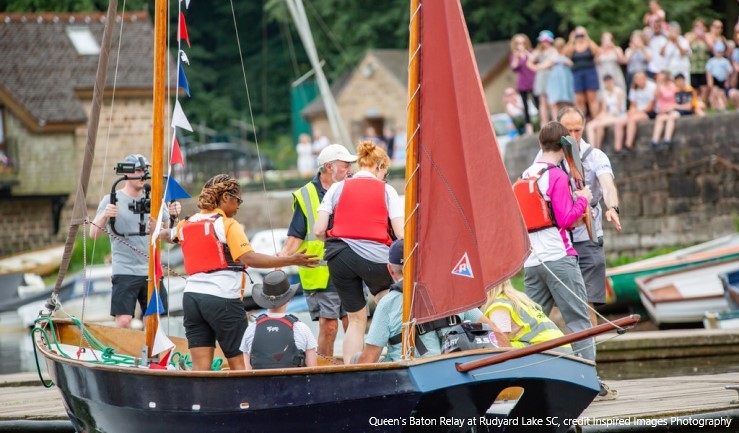 Queen's Baton Relay entourage disembarking a traditional boat at Rudyard Lake SC to welcoming crowds and with media following.
