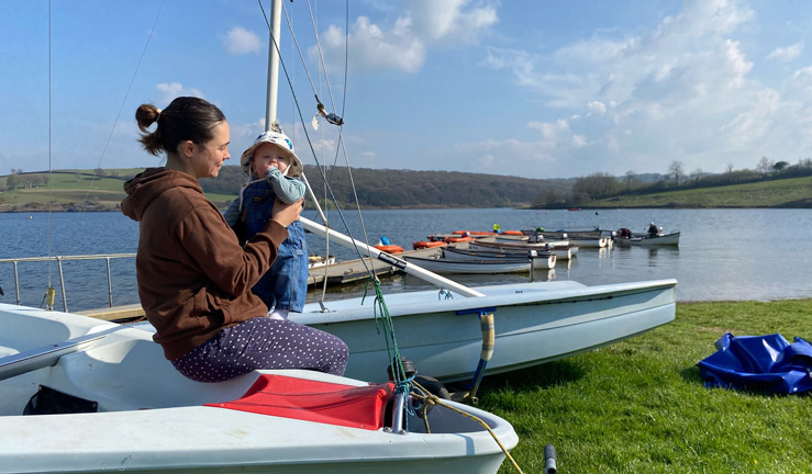Woman and her young daughter sat on side of boat in boat park looking out over boats moored up to pontoon on lake