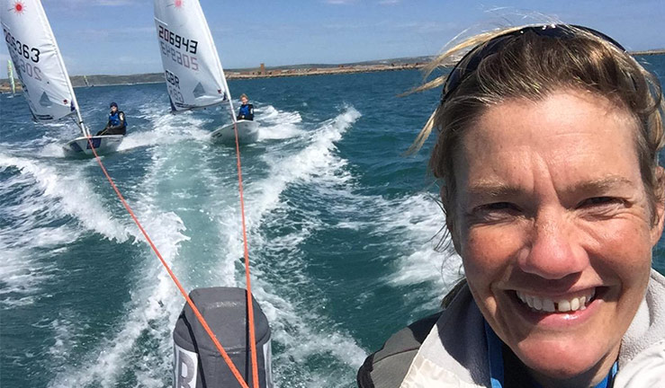 Penny Clark coaching. Penny is smiling at the camera, driving a RIB towing two dinghies behind. 