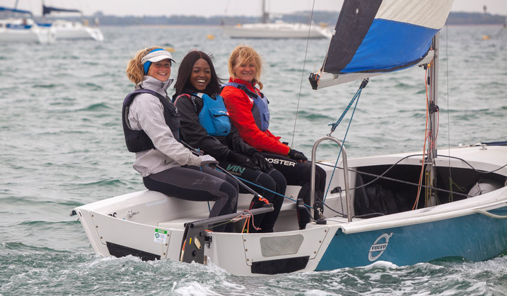 3 women sat in dinghy sailing. The boat is flat and they are all looking and smiling at the camera. There are yachts moored in the background. 