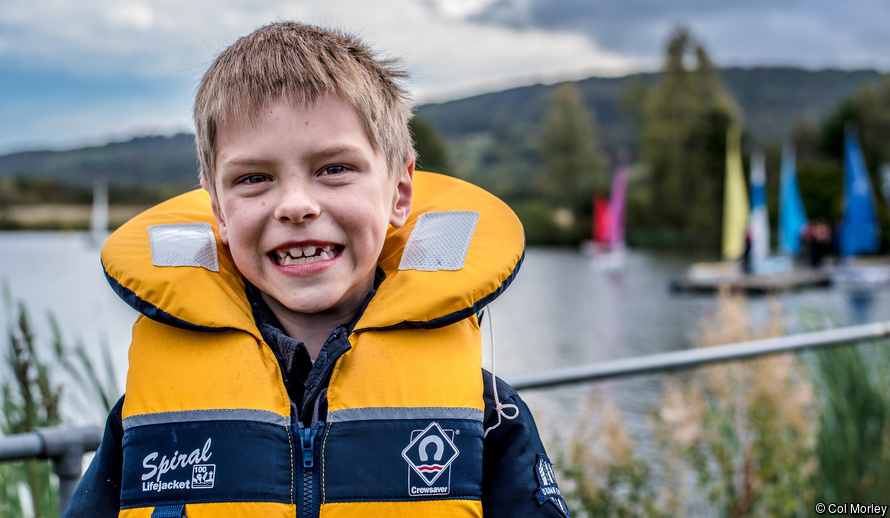 A young man, dressed for sailing with a huge smile, with a beautiful lake behind him with sailing boats