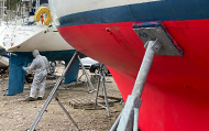 A boatyard with a red painted hull and a man in protective coating applying Anti-foul paint.