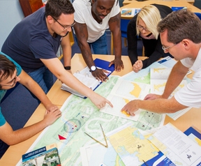 An image showing five people around a table in a classroom environment. They are looking at a paper chart on the table.