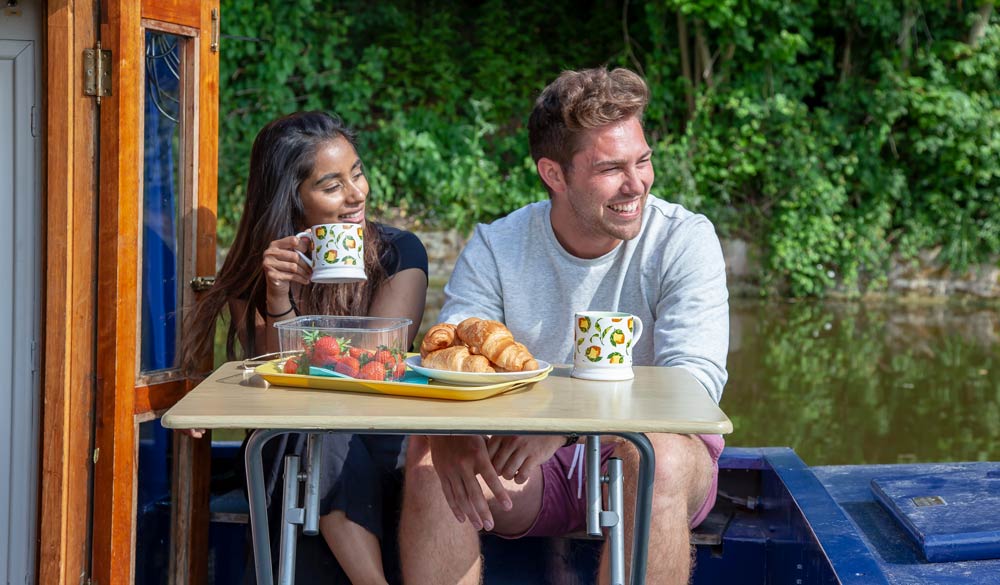 Man and woman sat at table at front of canal boat having breakfast
