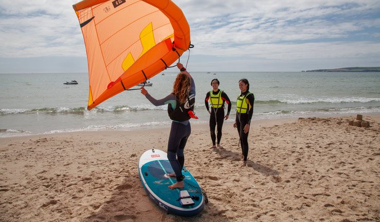 Learn to wingsurf lesson. Female instructor demonstrating with board and wing on sandy beach. Two students watching