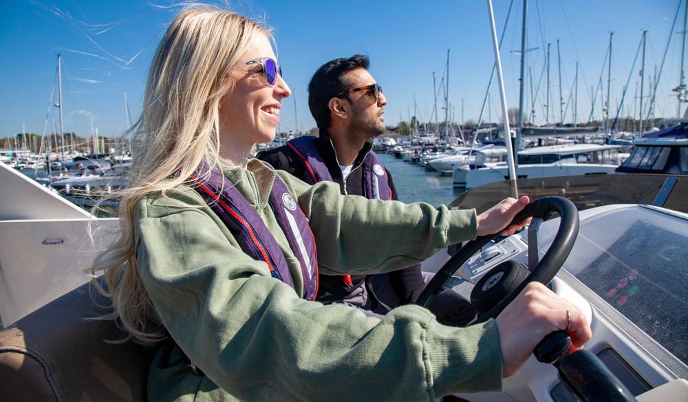 Woman helming motor cruiser looking ahead and smiling, man is seen in background sat next to her 