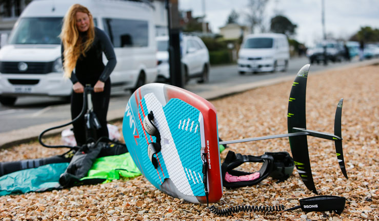 Woman inflating wing on beach. Wingfoiling board is visible in foreground.  