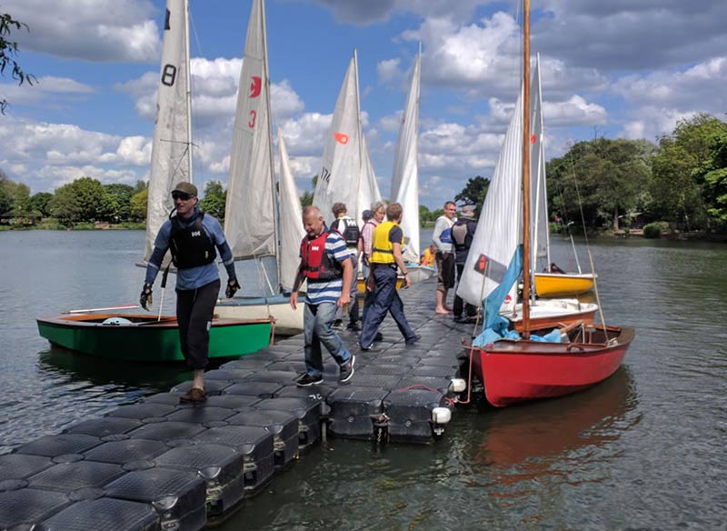 a fleet of sailing dinghies moored next to a temporary dock with a crowd of club members