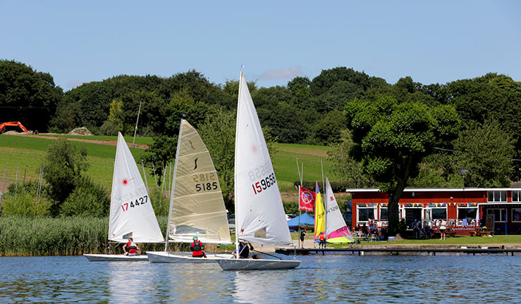 wide shot of sailing dinghies on a lake next to a club house on a sunny day.