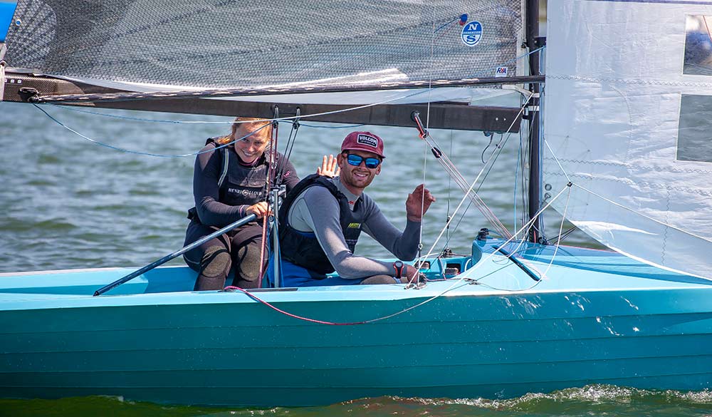 mid shot of a two people posing and waving at the camera sitting in a sailing boat