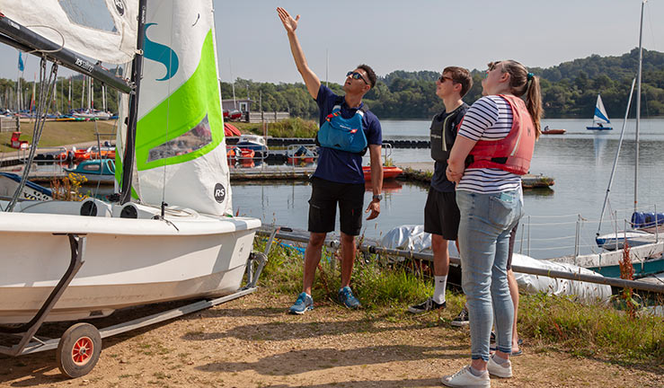 candid shot of an instructor talking to three teenagers as he points to a sailing dinghy  