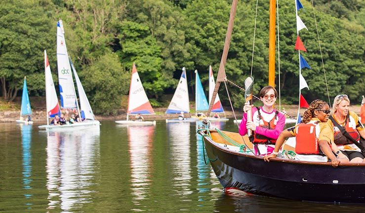 Queen's Baton Relay bearer for the Commonwealth Games 2022 pictured on a Drascombe Lugger crossing the water with other boats in the background at Rudyard Lake SC.