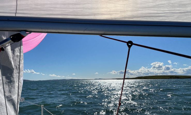 A view of the sea under the boom of a yacht. You can see the corner of a pink spinnaker and the sun is shining.
