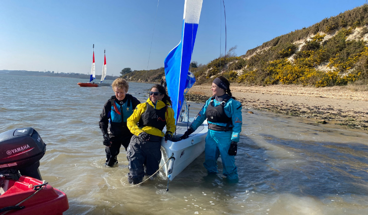 Three women in wetsuits, smiling and towing a dinghy in shallow water