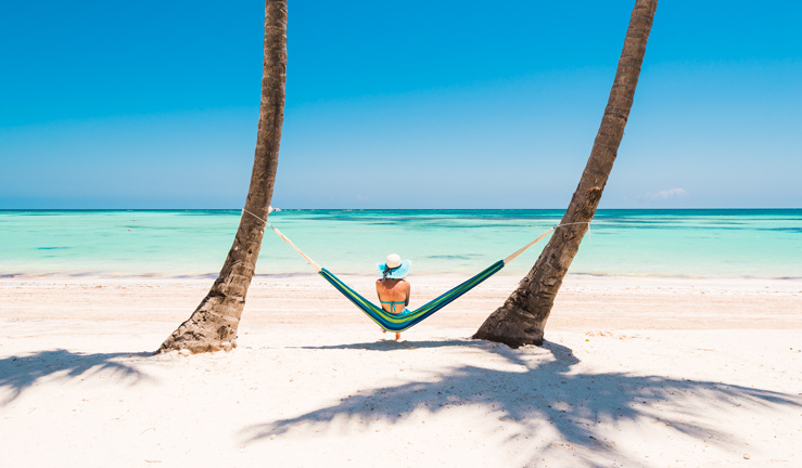 RYA Member reward partner offers September 2022 - P&O Cruises, women sat between palm trees on tropical, sandy beach looking out to sea. 