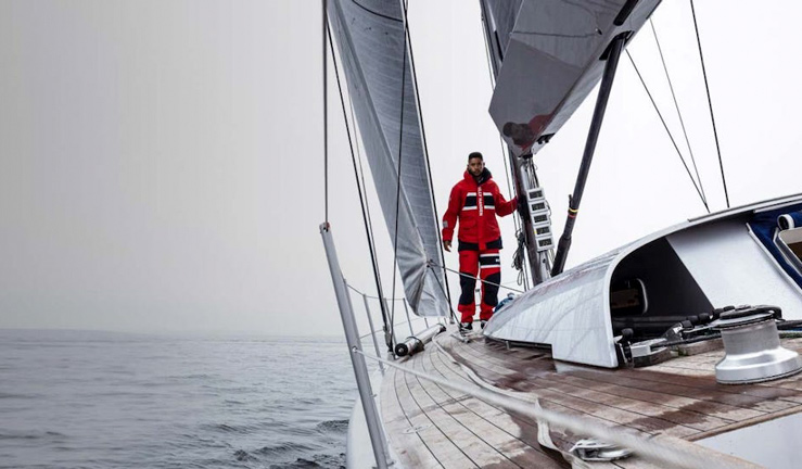 RYA member reward partners September 2022. Wetsuit Centre - man stood at bow of sailing yacht looking back towards cockpit wearing red Helly Hansen foul weather gear