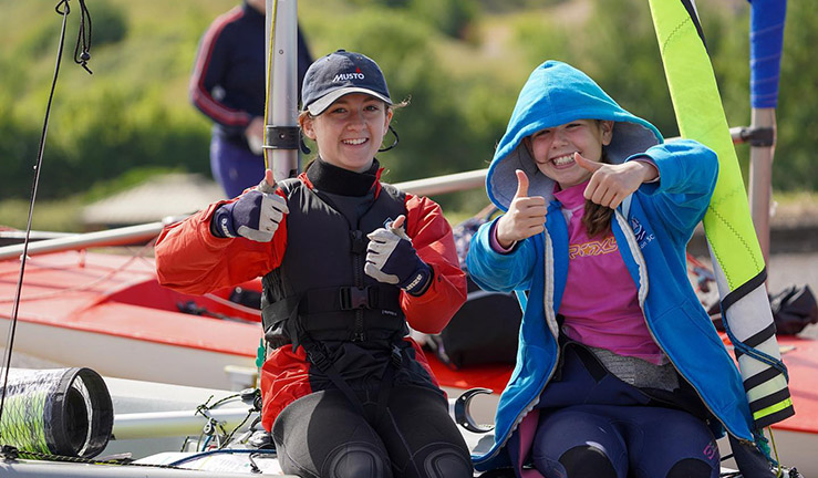 Two junior sailors sat on a boat on shore with smiles and thumbs up!