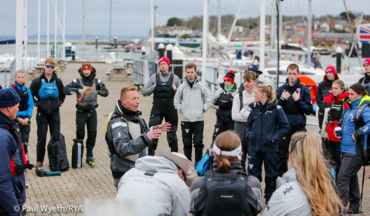 Group of British Keelboat Academy recruits on shore listening to a coach who is explaining something with his hands.