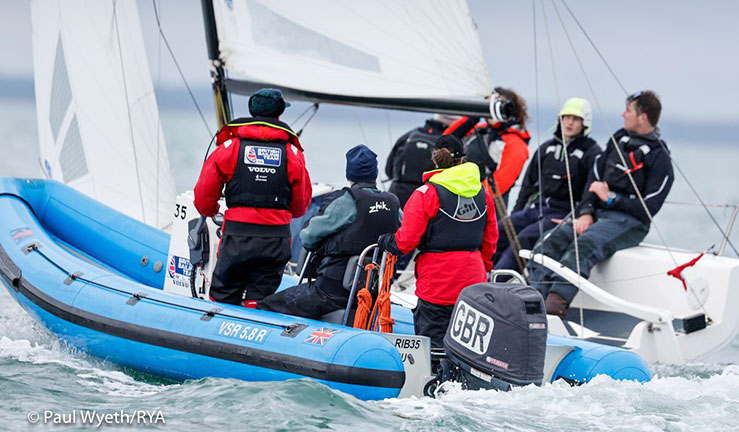 Blue RIB with coaches on board alongside a keelboat being sailed upwind by British Keelboat Academy recruits.