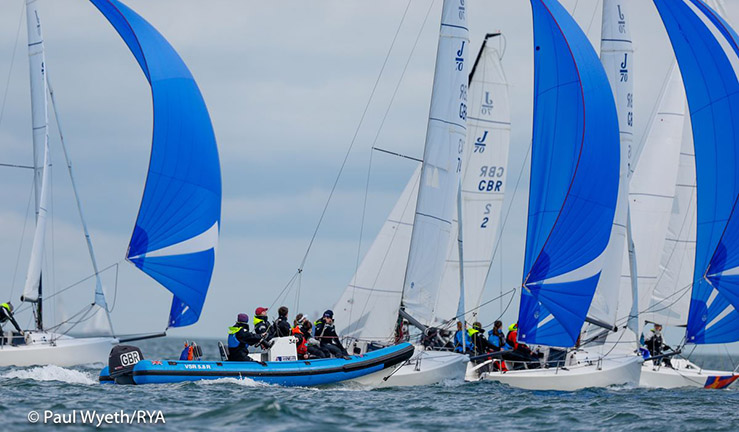 Coaches in a blue RIB alongside a fleet of J70 keelboats being sailed downwind by British Keelboat Academy recruits with blue spinnakers flying.