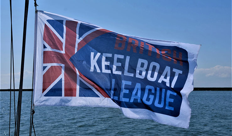 Red white and blue flag with British Keelboat League and union jack logo flying in the breeze with sky and sea in the background.