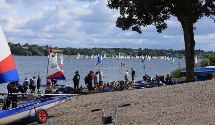 View from the shore at Draycote Water SC with junior boats on the water for the NSSA National Youth Regatta and people on shore with a couple of boats waiting to launch.