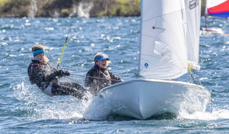 Two youth sailors racing a 420 dinghy on a sunny day with the crew trapezing, sparkling water and spray. 