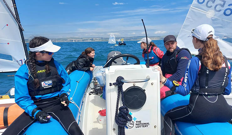 Optimist sailors gathered around blue coach boat on a sunny day at the national championships, Weymouth, 2023.
