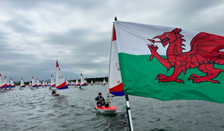 Welsh flag flying with Topper dinghy sailors on the water in the background at the national championships, Plas Heli, 2023