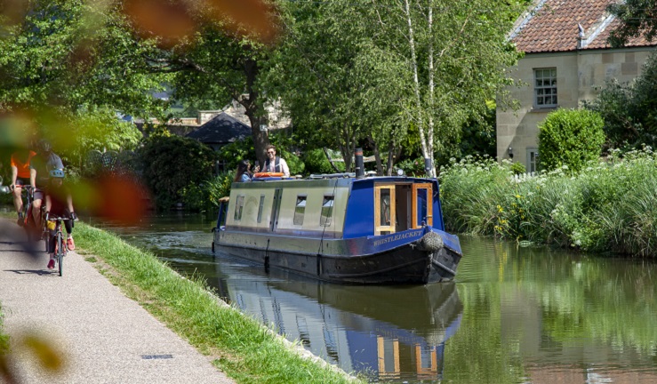 A narrowboat is travelling along a canal. Cyclists are riding along the towpath. 
