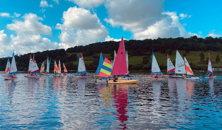 Around 20+ dinghies on Lake Bala with a hillside in the background, and with colourful sails and white clouds in a vibrant blue sky all reflecting on the water.