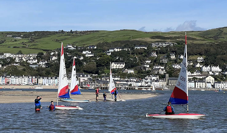Four junior Topper dinghies launching at Dovey Yacht Club from the beach on a sunny day with the town behind.