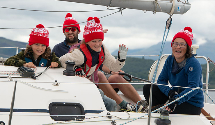 A smiling family in matching bright red hats sailing onboard a cruising yacht taking part In West Highland Yachting Week
