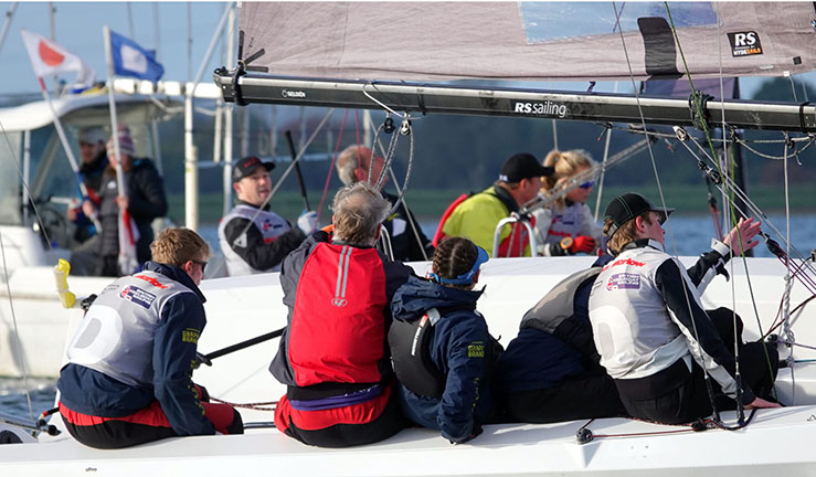 Close up view of two teams coming off the startline in RS21s with committee boat in the background at RYA National Match Racing Grand Finals 2023