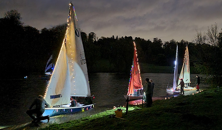 A night time picture of four boats lined up along the shore with people alongside in winter coats and hats at Himley Hall Sailing Club - all of the boats are lit up in the dark by fairy lights on their hulls, masts, sails and shrouds! 