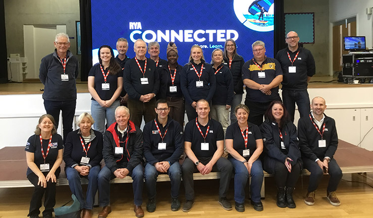 Team picture of RYA Midlands and head office staff, contractors and regional volunteers at RYA Connected event for the region at Brockington College, Nov 2023  - around 20 people on the stage with RYA Connected graphic on big screen behind.