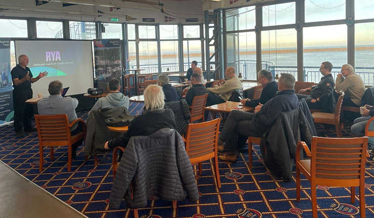 Group of people sat listening to a presentation at Sunderland Yacht Club at the RYA Connected North East Club Gathering, in a room with a view of the sea from the floor to ceiling height windows. 