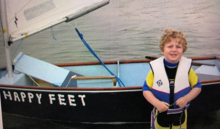 A child with blond curly hair, wearing a buoyancy aid and wetsuit, standing next to a dinghy called Happy Feet