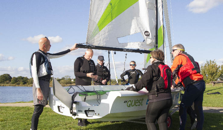 Sailors stand around an RS Quest dinghy where an instructor is explaining points of theory 