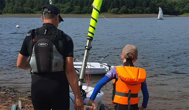over the shoulder shot of man and young child wearing life preservers pulling sailing dinghy into lake