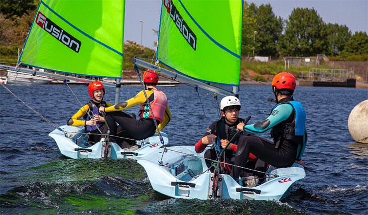 Two Fusion dinghies on a sunny day each with two youth sailors on board, one helming, one crewing, wearing helmets.