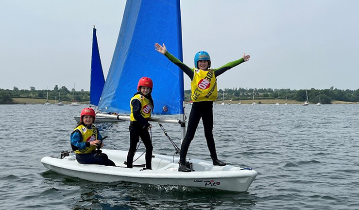 Three OnBoard sailors on a sailing dinghy, with the one on bow with his arms in the air smiling.