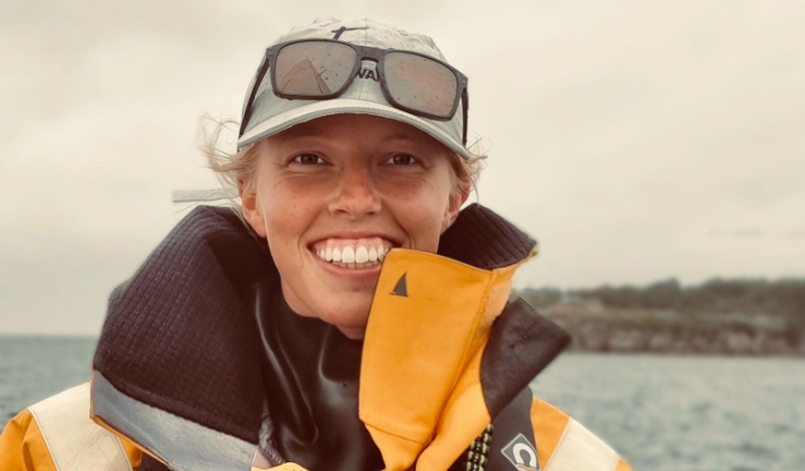 Meg wearing yellow dry suit and a baseball cap with sunglasses on top, smiling.