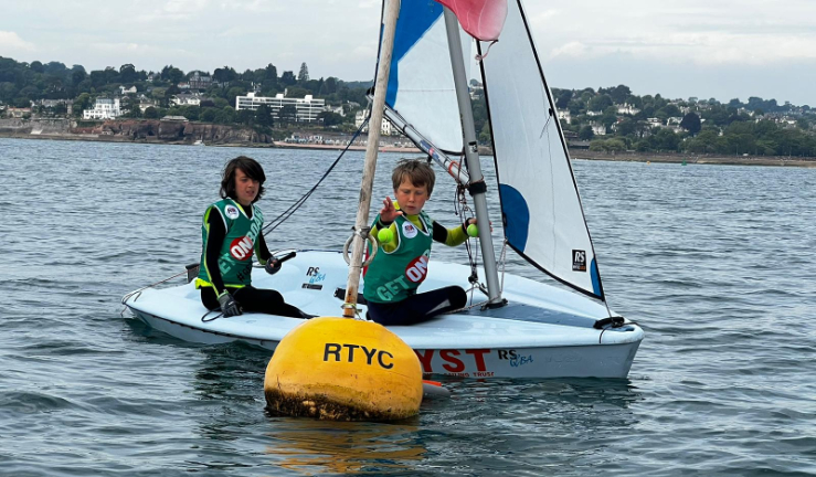 Two youngsters wearing OnBoard buoyancy aids in a dinghy and throwing tennis balls into a bucket attached to a buoy