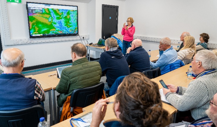 Penny Tranter presents a workshop to delegates sitting at tables, with a weather map on the screen.