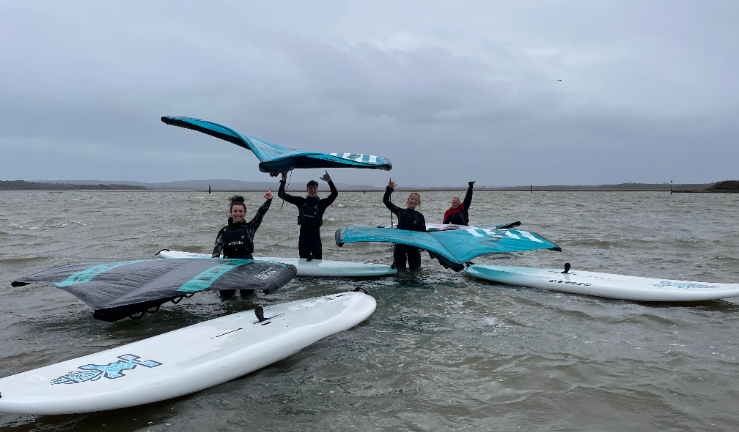 Four wingsurfers standing in shallow water with their kit, waving