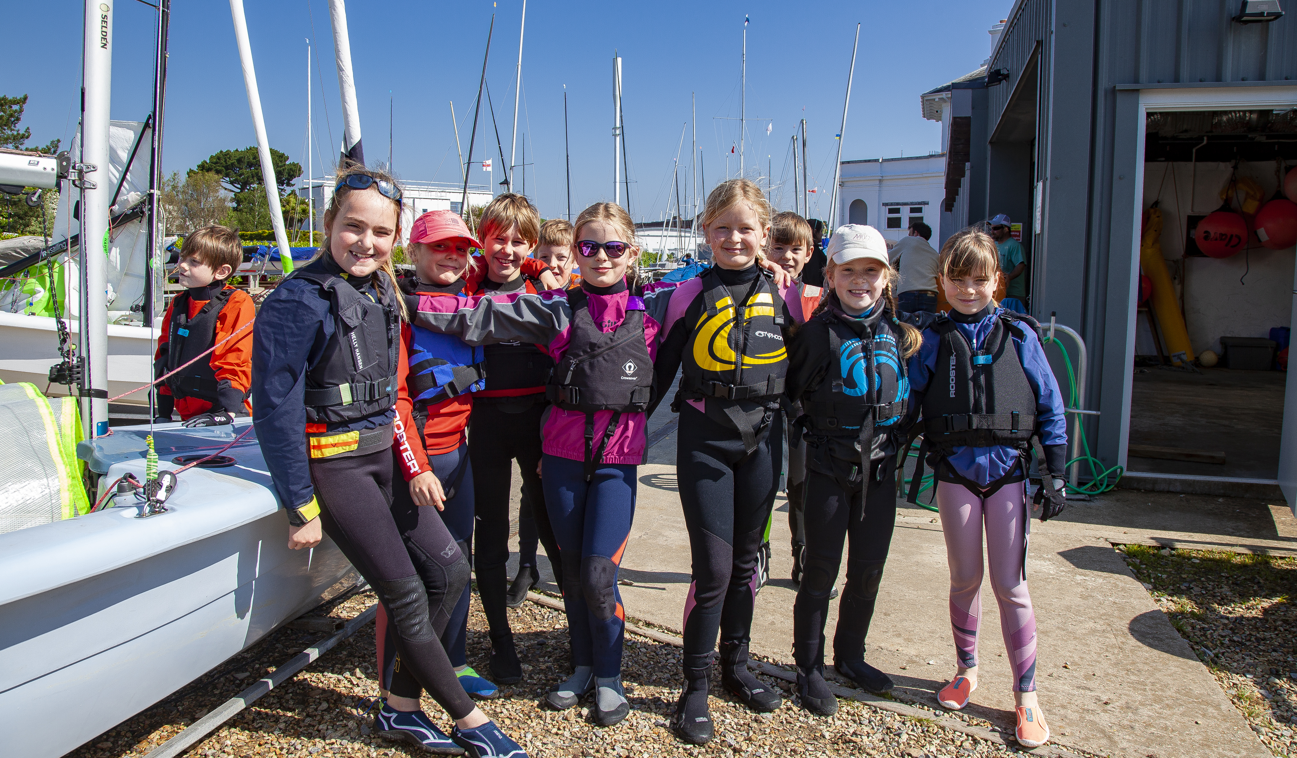 Children in sailing gear standing in a group next to a boat smiling. 