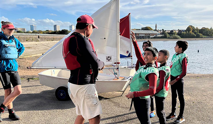 Group of four young sailors in OnBoard bibs chatting to an instructor on shore.