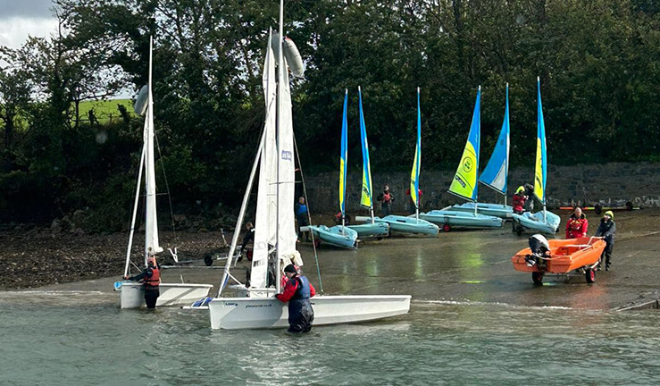Two dinghies and a RIB launching from the shore at Plas Menai with six training boats with blue and green sails on the slipway in the background.