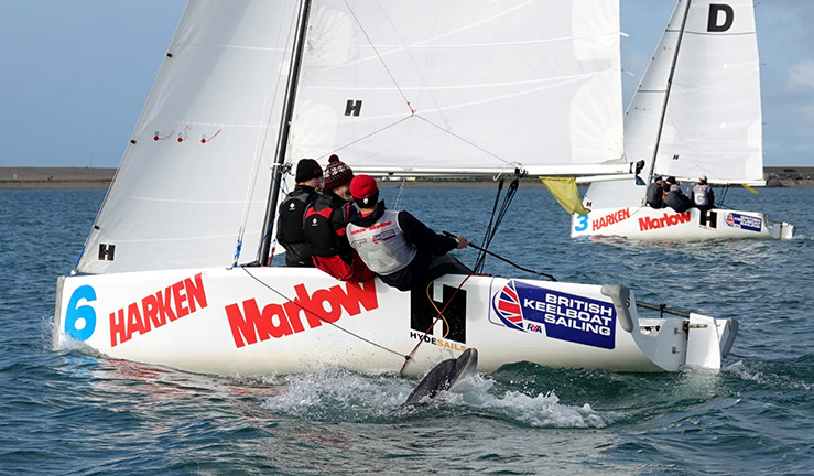 Two teams match racing upwind in Elliot 6m keelboats on a sunny day at WPNSA for the RYA Schools Match Racing Championship - with a dolphin just popping up by the side of the boat nearest the camera!